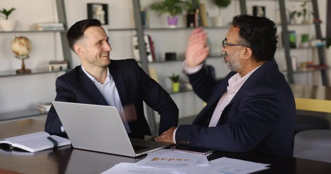 Two businessmen discuss joint project on laptop, giving high five gesture, symbolize celebrating achieving consensus on mutual decision, successfully overcoming challenge, finalizing a strategic plan