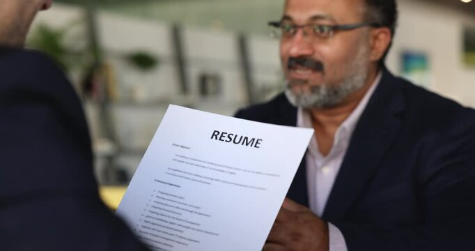 Close up of laughing male company position applicant answers questions during job interview meeting with HR, headhunter holds resume asks about education and qualification to mature vacancy candidate