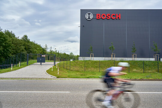 Large BOSCH building, cyclist in motion blur passing by, paved road leading to a gated entrance, depicting industrial architecture and motion. Wernau, Germany, Europe.