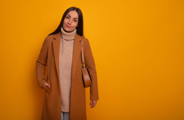 Lovely stylish woman wearing a camel coat, beige turtleneck sweater, and carrying a brown bag smiles softly against a vibrant yellow background. The look is warm, modern, and casual chic.