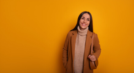 A smiling woman in a camel coat, beige turtleneck sweater, and carrying a brown bag stands against a vibrant yellow background. She exudes warmth, style, and confidence in a modern look.