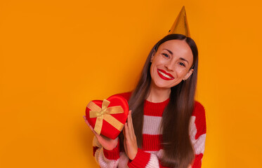 A joyful woman in a striped sweater and party hat holds a heart-shaped gift box with a golden ribbon, smiling brightly against an orange background. Perfect for festive and celebration themes.