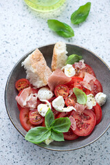 Plate of caprese served with torn prosciutto and fresh ciabatta, vertical shot on a light-grey granite surface, elevated view