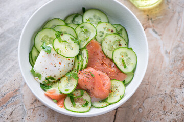 Bowl of cucumber salad with smoked salmon and poached egg, horizontal shot on a grey and roseate granite background