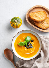 Pumpkin soup with cream, basil and seeds on a light background with bread, spoon and napkin.