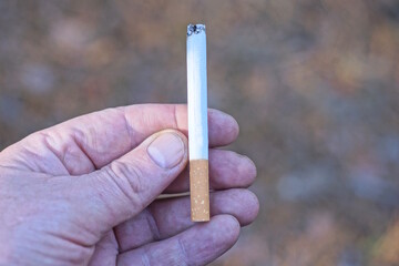 fingers on the hand hold one white cigarette with a brown filter outdoors on a gray background