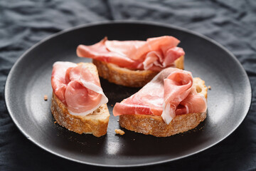 ciabatta with thin prosciutto on black plate on linen background