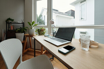 Contemporary home office space with open window view, showcasing laptop on wooden desk, smartphone beside. A well-organized, serene setting with greenery and natural elements