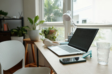 Minimalist home office featuring laptop on wooden desk, surrounded by lush green plants and natural light from window for a refreshing work environment