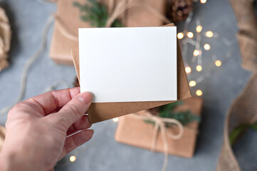 Obraz premium Christmas blank greeting card, postcard or invitation mockup, empty paper card in hand, blurred garland lights and gift boxes on background