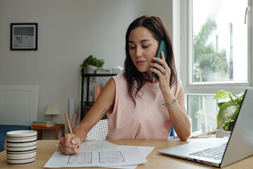Woman multitasking by discussing project plans on phone while examining blueprints at home office...