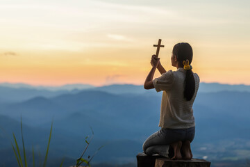 Girl holding Christian cross kneeling down praying to God on the mountain sunset background.