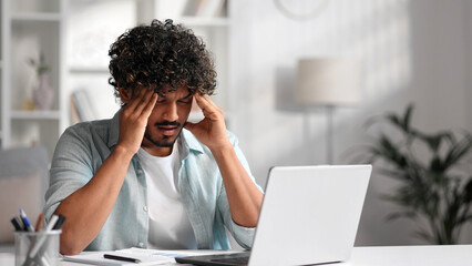 A young Indian male businessman is sitting at a desk trying to work, checking schedules and getting frustrated that something is not working out. Headache, fatigue, burnout.