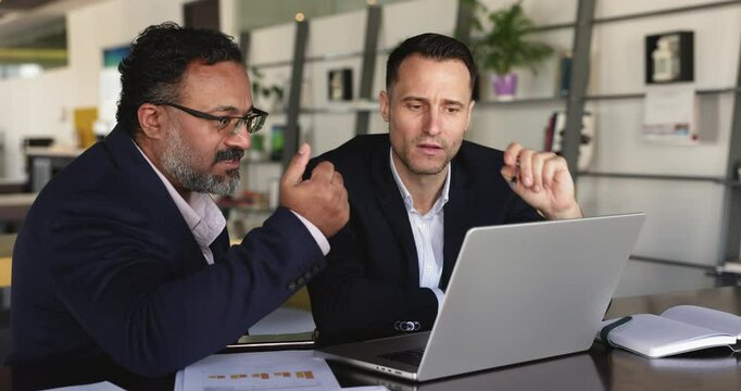 Two business men engaged in on-line project discussion, sit at desk with laptop, analyzing diagrams and charts, review trends, exchanging opinions, learn data, considering various strategic options