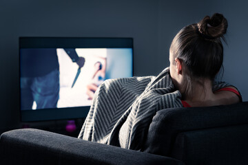 Watching horror and scary film on tv. Woman and thriller movie on television. Person in fear on home sofa couch under blanket at night in dark. Streaming true crime series or halloween show.