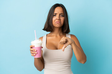 Young woman with strawberry milkshake isolated on blue background showing thumb down with negative...