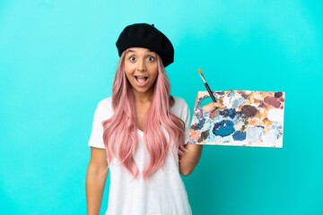 Young mixed race woman with pink hair holding a palette isolated on blue background with surprise...