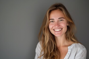 Obraz premium A smiling woman with long hair stands in front of a plain gray wall, exuding confidence, simplicity, and natural beauty in an indoor setting.