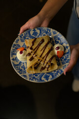 A woman showcases a decorative plate with perfectly folded crepes, drizzled with chocolate and garnished with whipped cream and fresh berries. The focus is on the dessert and its artistic presentation