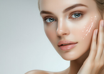Obraz premium A young woman with radiant skin applies a skincare product on her cheek, shown in smooth strokes. She gently touches her face, highlighting the hydration and glow of her complexion in a clean setting