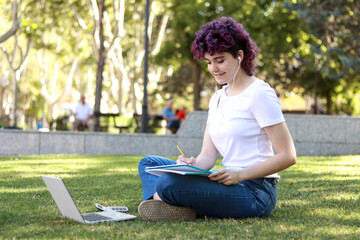 Young student with purple curly hair sitting on the grass with a notebook and laptop studying in the park