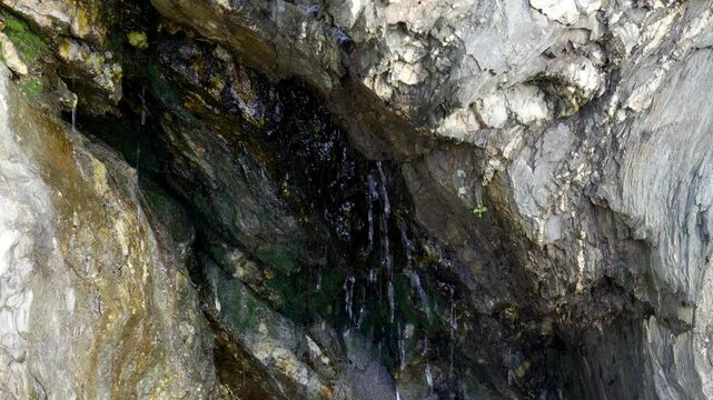 A close-up view of a rocky cliff face with a small stream of water trickling down. The rock is dark grey and brown with patches of green moss.