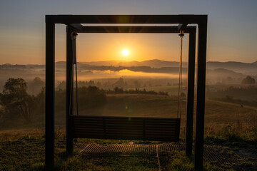 Sunrise over misty hills with mountains in the background framing a large wooden swing