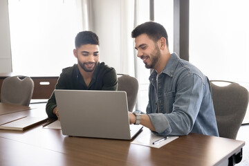 Obraz premium Two positive young male Middle Eastern entrepreneurs working on Internet project, online startup together, watching website media content, pointing at monitor, smiling, sitting at table together