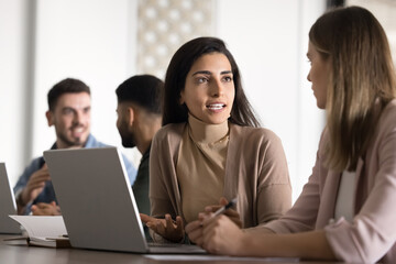 Confident Hispanic businesswoman in casual talking to female colleague at laptop, discussing online startup, teamwork plan, project solutions. Diverse employees working at computer together