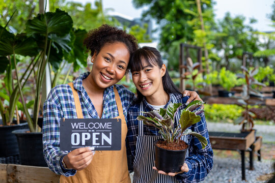 African gardener and owner with her asian partner are holding open sign in front of their ornamental houseplant at their own nursery garden center full of exotic and tropical plants