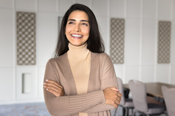 Happy dreamy confident Latin business professional woman standing with arms crossed in office workspace, looking away with toothy smile, thinking on successful career. Businesswoman casual portrait