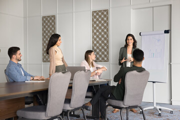 Confident positive young Latin business trainer woman presenting project statistic data to diverse...