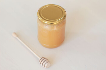 A minimalist still life photograph showcasing a glass jar filled with golden honey and a wooden honey dipper. The light background enhances the warm tones of the honey. Stock photo