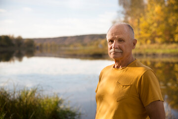 Obraz premium portrait smiling senior man in a yellow T-shirt walking on the shore of the lake on a warm autumn day. copy space.