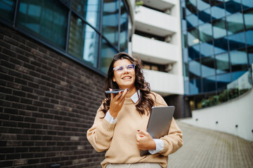 Young businesswoman sending voice message in front of modern business building	
