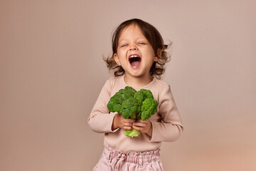 smiling child girl holding fresh green raw broccoli on beige background. healthy baby food. kid loves vegetables