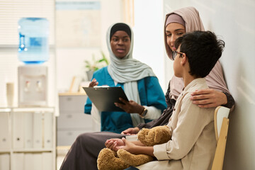 Group of caring Muslim professionals and child with teddy bear sitting in modern medical office, showing support and comfort during healthcare visit, affectionate interaction, diverse group