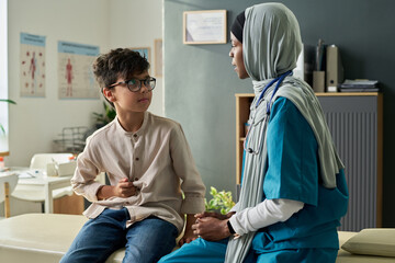 Caring Muslim physician wearing hijab and scrubs sits beside young boy, providing comfort and...