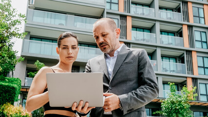 A businessman and a young female assistant discuss a new project outdoors while using a laptop. Their collaboration highlights a dynamic and modern approach to work.