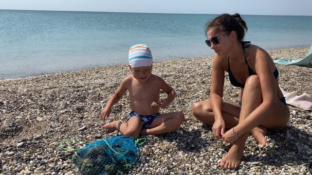 Woman and child look at fresh catch of rapa shells in a net. Mother with little boy enjoying beach holiday on vacation. Evpatoria, Crimea. Rapana venosa