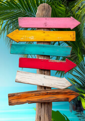 Colorful beach sign, wooden arrow signs pointing in various directions against a tropical background with palm trees and a beach.