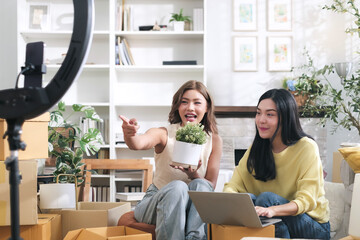 Beautiful asian woman and her friend showing off products while live selling online using phone and laptop.
