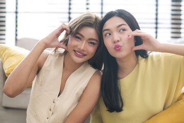 Portrait two young asian woman happy smile showing heart hands touch cheek gesture on sofa at home.
