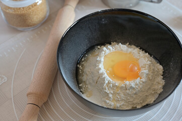 Baking ingredients for baking sweet buns, close-up flour with a broken egg in a black bowl, rolling pin on a table