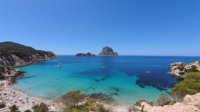 Stunning panorama of Ibiza Cala Hort beach, with Es Vedra and Es Vedranell islands visible in the distance Yachts float on the clear waters, while beachgoers relax on the sandy shore