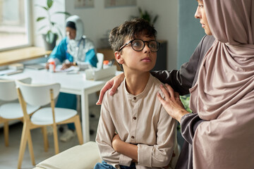 Obraz premium Muslim woman gently reassuring her son in warm, inviting living room as another family member stays in background at a dining table, creating a peaceful atmosphere