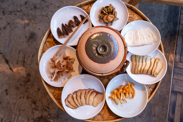 A close-up shot of various types of mushrooms and tofu slices arranged on white ceramic plates, placed around a traditional clay pot on a bamboo tray. 