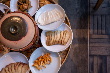 A close-up shot of various types of mushrooms and tofu slices arranged on white ceramic plates, placed around a traditional clay pot on a bamboo tray. 