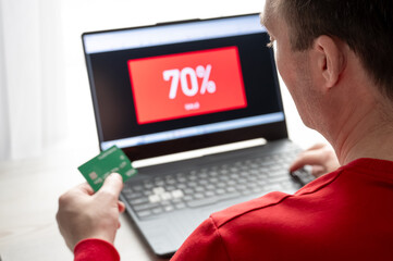 Man holding a credit card while shopping online on a laptop showing a 70% discount sale screen. The...