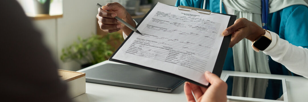 Medical professional holding clipboard with patient's records, examining details. Tablet device visible on desk during consultation in modern clinical setting - Powered by Adobe